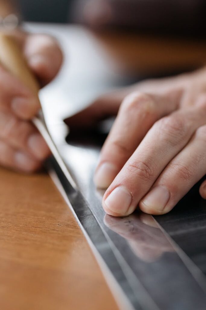 Hands carefully marking and cutting with a ruler and knife on a reflective surface.